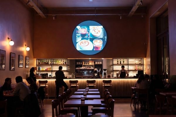 A modern café interior with a large round LCD display module on the wall showing food and drink animations