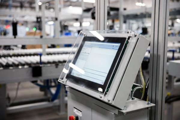 Large 11-inch LCD display module embedded in a rugged operator console on a logistics sorting line, showing a high-contrast sorting UI under warehouse lighting.