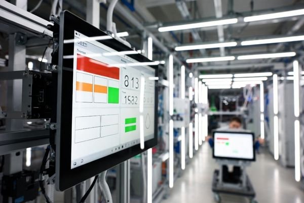 Wall-mounted production line dashboard LCD display module viewed off-axis on a factory floor, showing simple red/amber/green status blocks and a maintenance cart module for replacement.