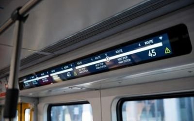Ultra-wide bar-type LCD module mounted above a train carriage door, showing a high-contrast passenger information layout under cabin lighting.