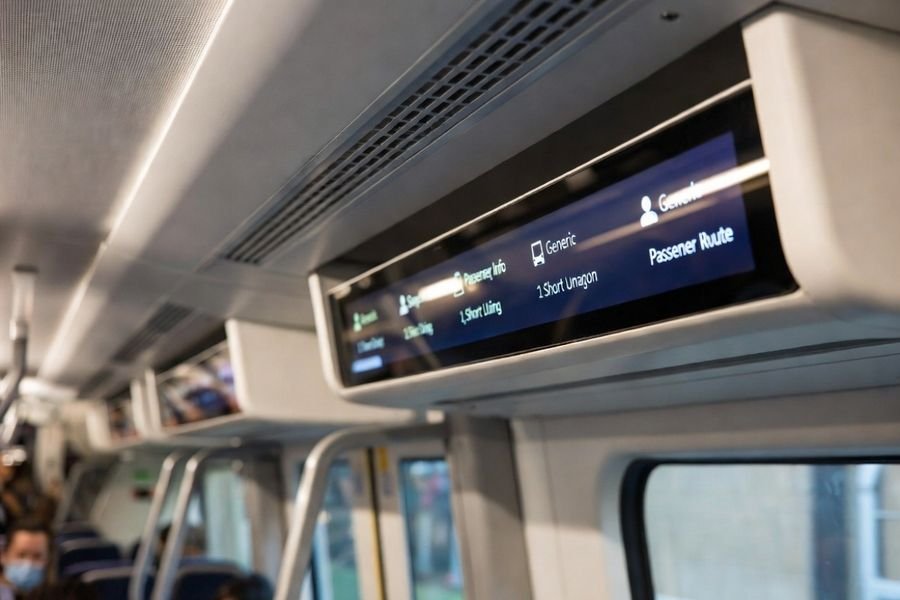 Ultra-wide bar-type LCD module mounted above a train carriage door, showing a high-contrast passenger information layout under cabin lighting.
