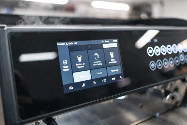 Commercial coffee machine panel with integrated LCD display module and touch interface in a bright café environment