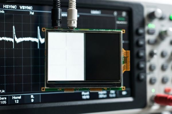 Realistic lab setup showing an LCD display module with a shifted border test pattern and an oscilloscope verifying sync timing signals.