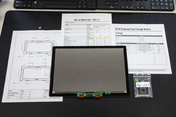 A realistic sample approval documentation review scene showing an LCD module, datasheet, mechanical drawing, BOM version note, firmware label, inspection report, and change record checklist on an engineering desk.