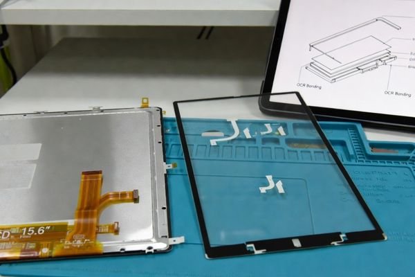 A realistic front-surface stack review showing an LCD module, PCAP touch sensor, cover glass sample, bonding layer diagram, and touch controller board on an engineering workbench.
